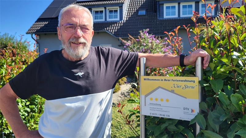 Älterer Mann mit Brille und Bart lehnt sich an ein Schild vor einem Haus mit Garten