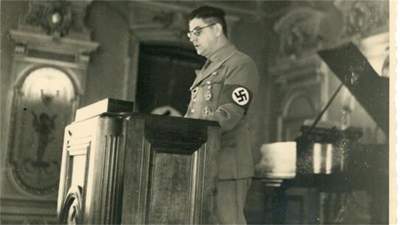 Mann in NS-Uniform mit Brille und Hakenkreuz-Armbinde steht an einem Rednerpult in einem prunkvollen Raum