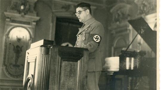 Mann in NS-Uniform mit Brille und Hakenkreuz-Armbinde steht an einem Rednerpult in einem prunkvollen Raum