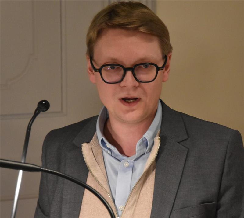 Mann in Sakko und hellblauem Hemd steht hinter einem Mikrofonständer