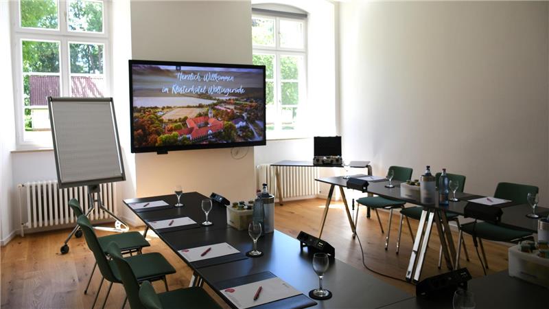 In einem Raum mit Holzfußboden stehen Tische und Stühle für Seminarteilnehmer bereit, an der Wand hängt ein großer Flachbildschirm.
