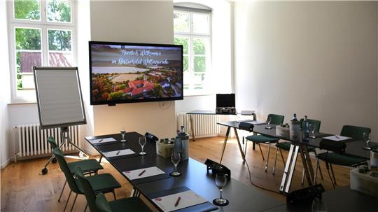 In einem Raum mit Holzfußboden stehen Tische und Stühle für Seminarteilnehmer bereit, an der Wand hängt ein großer Flachbildschirm.