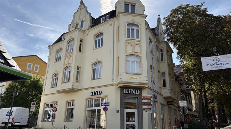 Eckgebäude mit mehreren Stockwerken und Ladengeschäft "KIND" im Erdgeschoss an einer sonnigen Straßenecke mit Bäumen und weiteren Gebäuden im Hintergrund