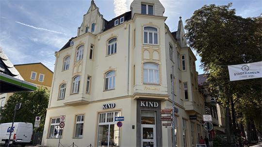Eckgebäude mit mehreren Stockwerken und Ladengeschäft "KIND" im Erdgeschoss an einer sonnigen Straßenecke mit Bäumen und weiteren Gebäuden im Hintergrund