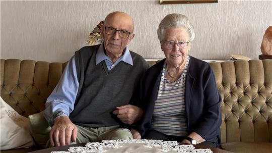 Älteres Paar sitzt eng nebeneinander auf einem braunen, gepolsterten Sofa mit Zierkissen und einer weißen Spitzen-Tischdecke im Vordergrund