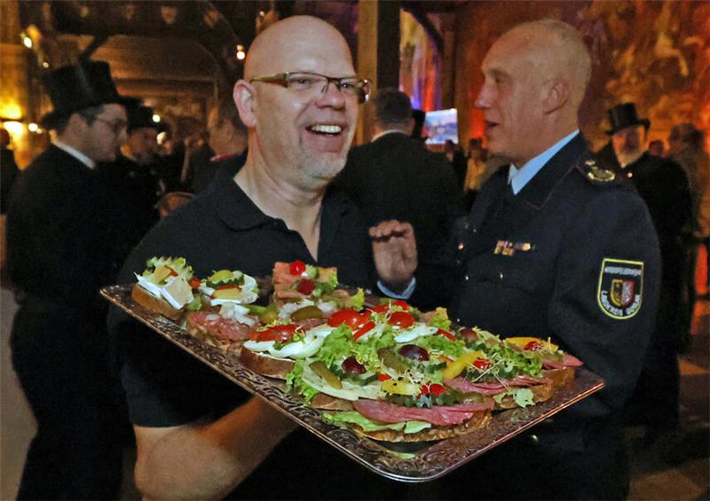 Zwei Männer, einer in Uniform, halten ein Tablett mit verschiedenen belegten Broten und Salatblättern in einer festlich beleuchteten Umgebung.