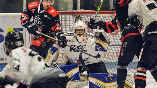 Eishockeyszene vor dem Tor mit Torwart in weiß-blauem Trikot und mehreren Spielern in schwarz-roten und weißen Trikots, die um den Puck kämpfen.