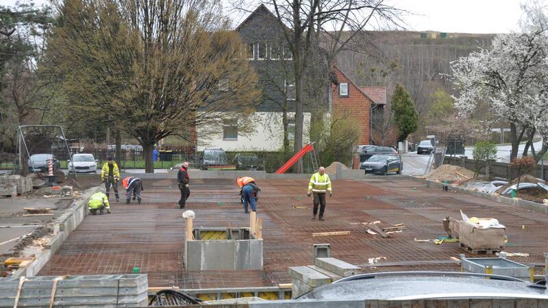 Mehrere Bauarbeiter verteilen sich auf einer Baustelle mit Betonboden und Bewehrungseisen vor Wohnhäusern und Bäumen