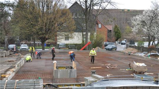Mehrere Bauarbeiter verteilen sich auf einer Baustelle mit Betonboden und Bewehrungseisen vor Wohnhäusern und Bäumen