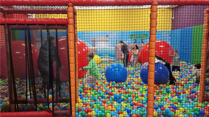 Kinder spielen in einem bunten Bällebad mit großen blauen Bällen und Klettergerüst im Hintergrund.