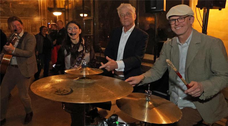 Mehrere Personen spielen in einem Innenraum Musikinstrumente, darunter Schlagzeug und Gitarre.