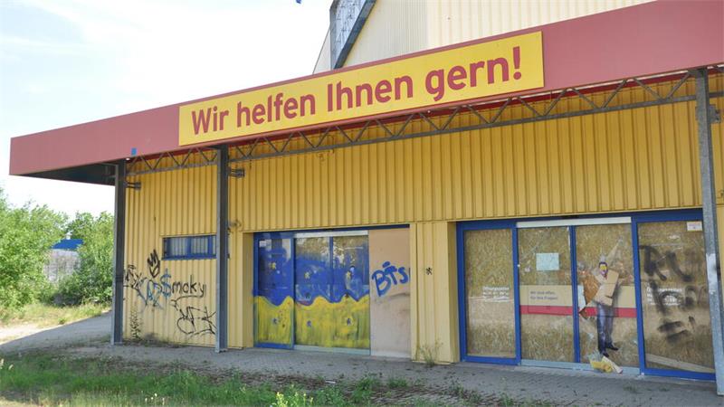 Verlassenes gelb-rotes Gebäude mit Graffiti an den Wänden und einem Schild mit der Aufschrift 'Wir helfen Ihnen gern!'