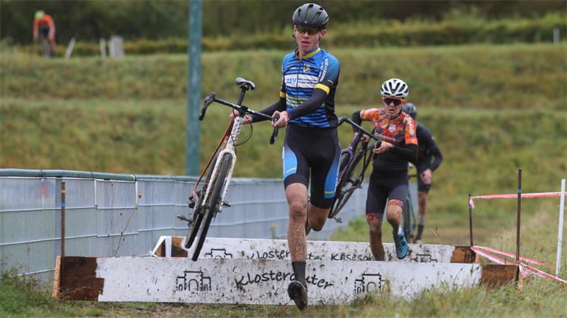 Zwei Radfahrer in Sportkleidung tragen ihre Fahrräder über eine Holzhürde auf einem schlammigen Weg