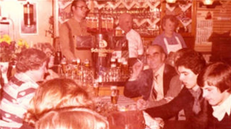 Menschen sitzen und stehen in einer Bar mit Holztheke und Regalen voller Flaschen im Hintergrund.