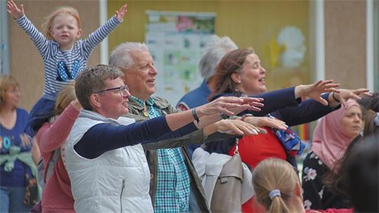 Mehrere Menschen unterschiedlichen Alters stehen mit ausgestreckten Armen in einem Innenraum, im Hintergrund ein Kind auf den Schultern einer Person