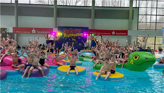 Viele Kinder und Jugendliche sitzen auf bunten Schwimmringen und einem grünen Schwimmtier im Hallenbad und heben die Arme