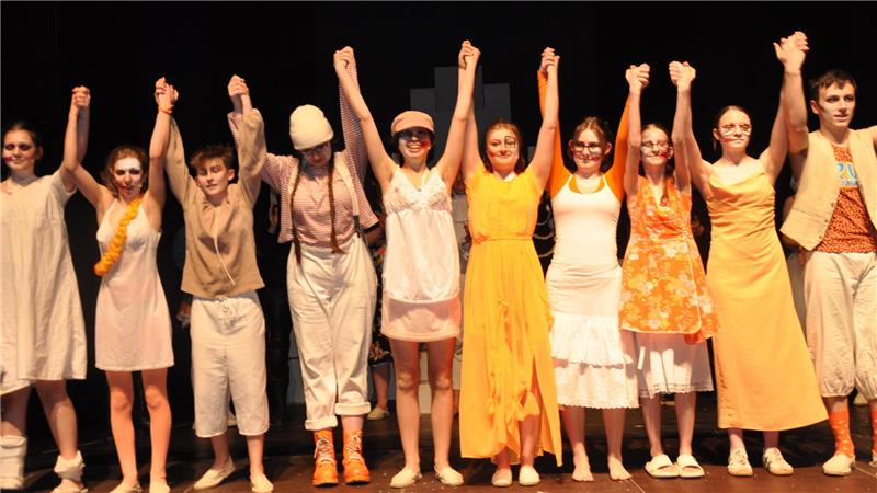 Gruppe von Menschen in bunter Kleidung steht auf Bühne und hält sich an den Händen hoch