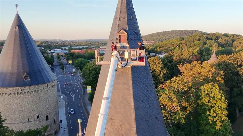 Zwei Personen auf einer Hebebühne an der Spitze eines steinernen Turms mit umliegenden Bäumen und einer Straße im Hintergrund