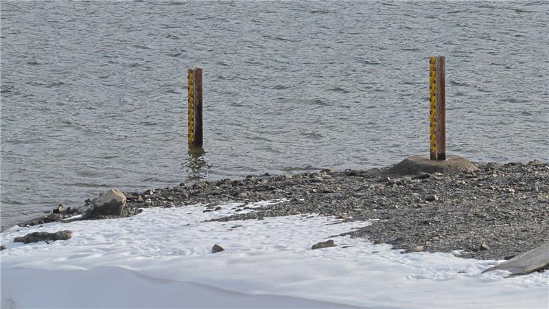 Zwei rostige Pfosten stehen im Wasser nahe einem kiesigen Ufer mit Schnee im Vordergrund.
