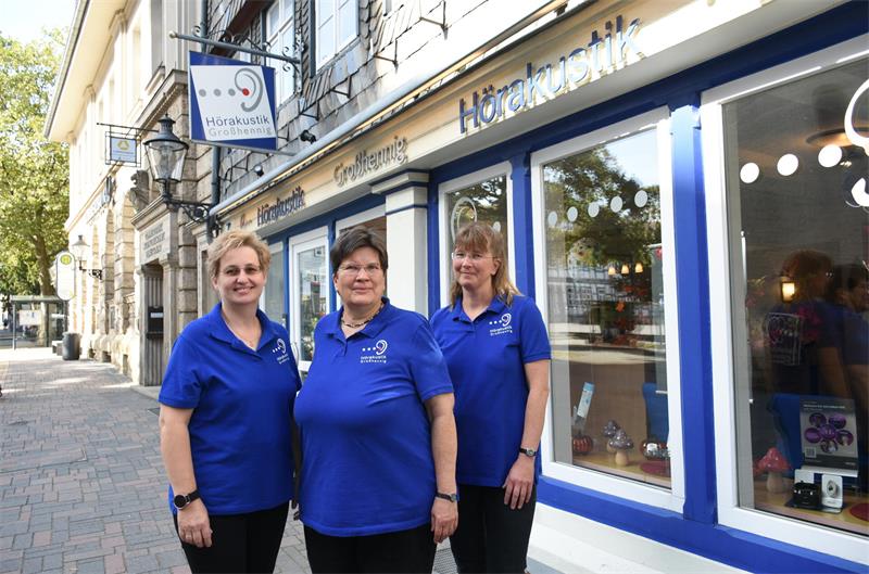 Drei Frauen in royalblauen Poloshirts stehen vor einem Ladengeschäft; über den Fenstern steht der Name "Großhennig".