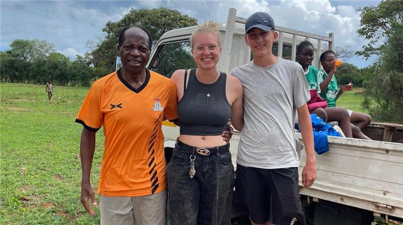 Drei Personen stehen nebeneinander auf einer Wiese vor einem weißen Pickup, im Hintergrund sitzen weitere Personen auf der Ladefläche.