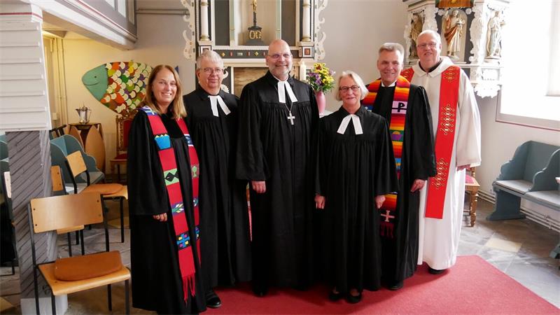 Sechs Geistliche in liturgischer Kleidung stehen in einer Kirche vor einem Altar mit Kreuz und Blumenstrauß.