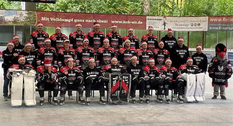 Gruppenfoto einer Eishockeymannschaft in schwarzen Trikots mit roten und weißen Streifen auf einer Eisbahn mit Werbebannern im Hintergrund