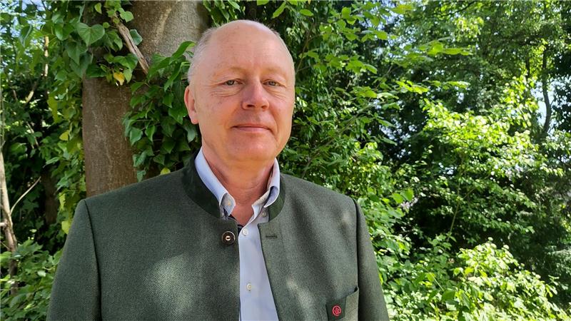 Mann mit grauem Sakko und hellblauem Hemd steht vor einem Baum und dichtem Grün
