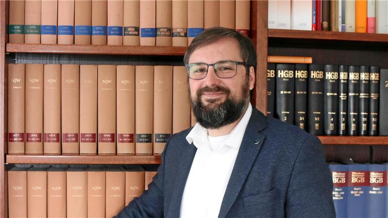 Ein Mann in Sakko und Hemd steht vor einem Bücherregal mit mehreren Reihen dicker, einheitlich gebundener Bücher und hat eine Hand auf einem Buch.