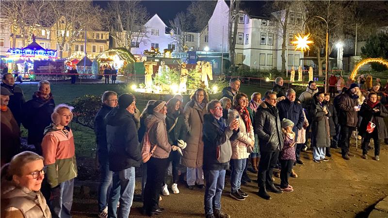 Viele Menschen versammeln sich vor der Weihnachtskrippe im Rosengarten, die im Hintergrund beleuchtet ist. 