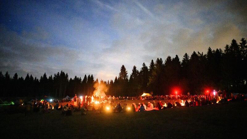 Hunderte Mitglieder der „Rainbow Family“ sitzen in einem großen Kreis rund um ein Lagerfeuer und essen das gemeinsam zubereitete Abendessen.