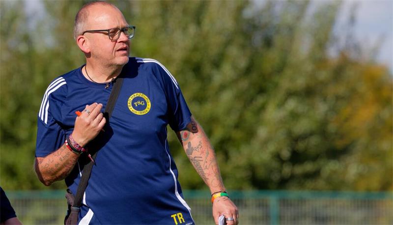 Mann mit Brille und Tattoos trägt blaues Sportshirt mit gelbem TSG-Logo und hält Stift und Notizblock auf einem Sportplatz.