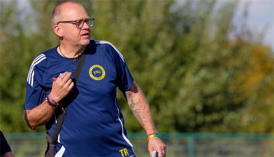 Mann mit Brille und Tattoos trägt blaues Sportshirt mit gelbem TSG-Logo und hält Stift und Notizblock auf einem Sportplatz.