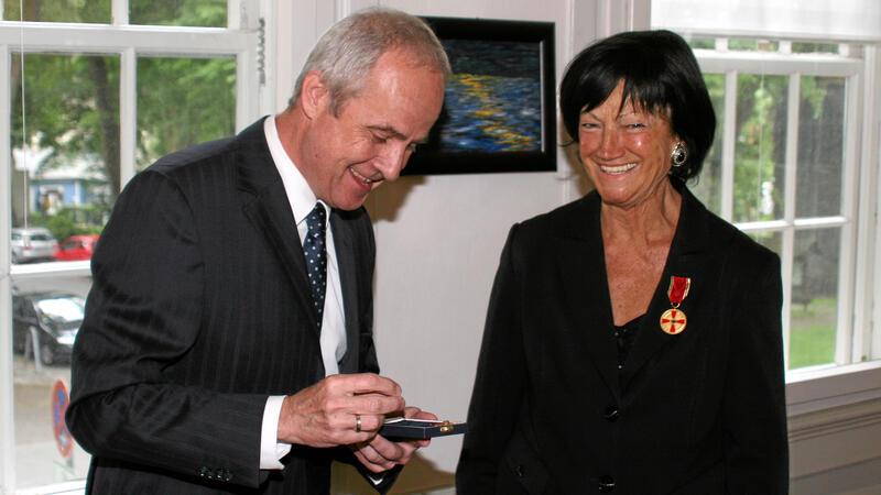 Mann im Anzug hält eine kleine Schatulle, während eine Frau mit Medaille am Revers daneben steht und lächelt.