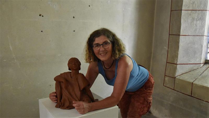 Frau mit Brille und lockigem Haar lehnt sich lächelnd an eine braune Skulptur auf einem weißen Podest in einem Kirchenraum mit hellem Mauerwerk und Fenster.