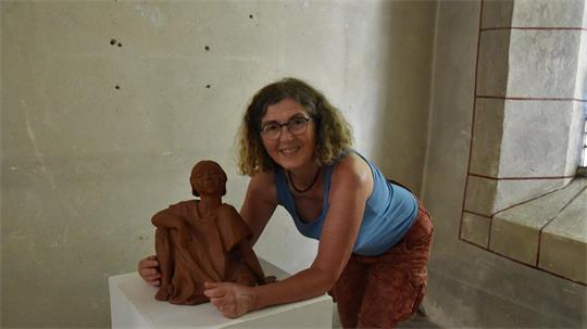 Frau mit Brille und lockigem Haar lehnt sich lächelnd an eine braune Skulptur auf einem weißen Podest in einem Kirchenraum mit hellem Mauerwerk und Fenster.