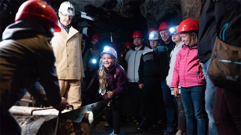 Gruppe von Kindern und Erwachsenen mit Schutzhelmen in einem dunklen Stollen, ein Kind sägt an einem Holzstück