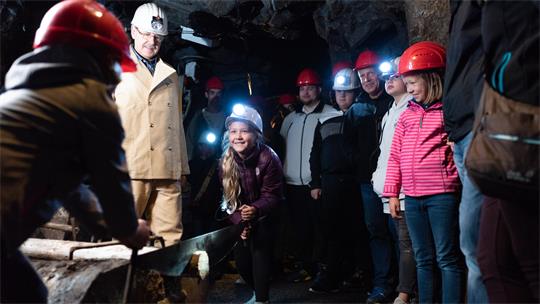 Gruppe von Kindern und Erwachsenen mit Schutzhelmen in einem dunklen Stollen, ein Kind sägt an einem Holzstück