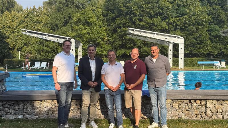Fünf Männer stehen nebeneinander vor einem Schwimmbecken mit Sprunganlagen und Bäumen im Hintergrund
