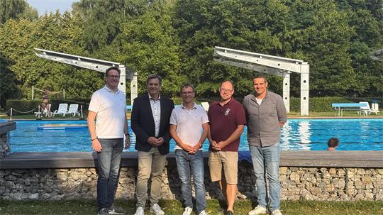 Fünf Männer stehen nebeneinander vor einem Schwimmbecken mit Sprunganlagen und Bäumen im Hintergrund