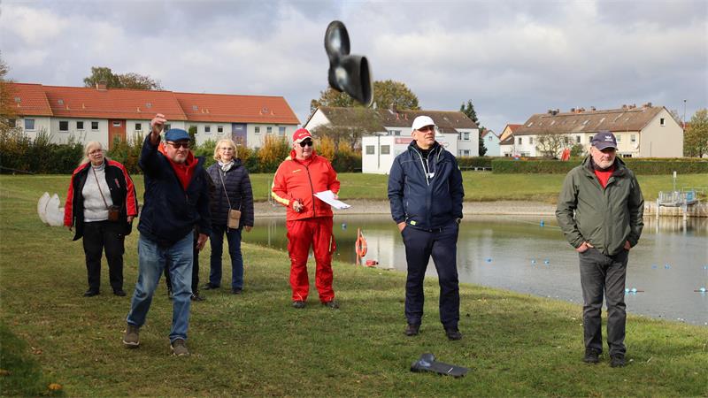 Gruppe von sechs Personen steht auf einer Wiese neben einem Teich, eine Person wirft einen schwarzen Hut in die Luft