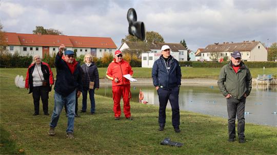 Gruppe von sechs Personen steht auf einer Wiese neben einem Teich, eine Person wirft einen schwarzen Hut in die Luft