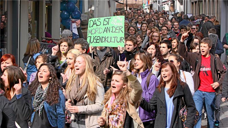 Menschenmenge bei einer Demonstration auf einer Straße, eine Person hält ein grünes Schild mit der Aufschrift 'DAS IST MORD AN DER JUGENDKULTUR'