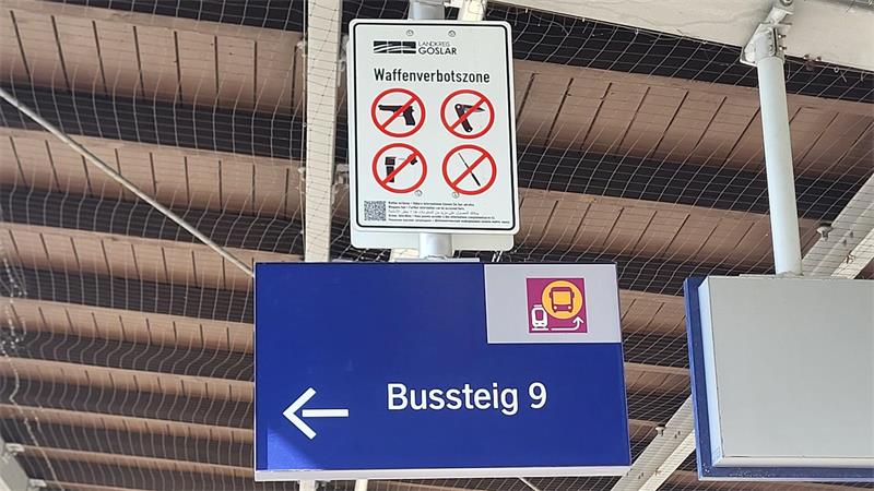 Schild mit Waffenverbotssymbolen über blauem Schild mit weißer Aufschrift 'Bussteig 9' und Pfeil nach links unter einem Dach mit sichtbaren Holzbalken