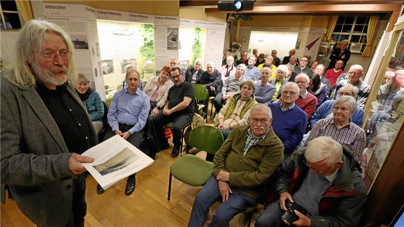 Ein Mann mit einer Broschüre in der Hand hält einen Vortrag. Dutzende Menschen hören zu.