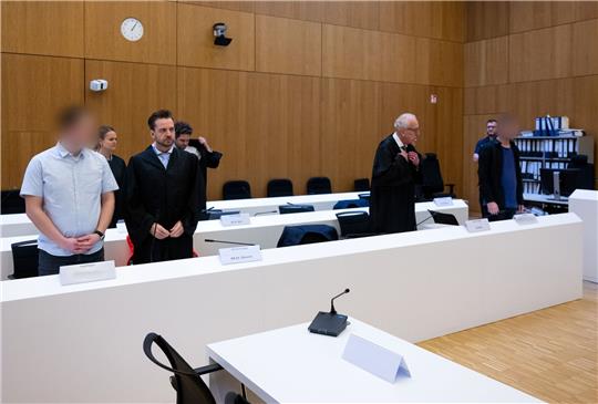 Im Hochsicherheitsgerichtssaal vom Landgericht München II stehen die zwei wegen Mordes angeklagten Männer (l und r) neben ihren Rechtsanwälten.