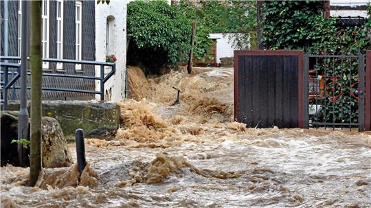 Überflutete Straße mit braunem, schäumendem Wasser, das zwischen Häusern und Zäunen hindurchfließt