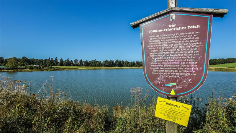 Schild mit Text zum Johann-Friedricher-Teich vor dem Teich und Wiesen im Hintergrund