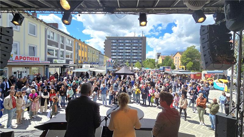Blick von einer Bühne auf eine große Menschenmenge bei einer Veranstaltung auf einem Marktplatz mit mehrstöckigen Gebäuden und blauem Himmel mit Wolken