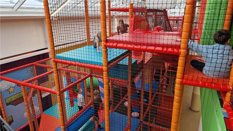 Mehrstöckige Kletteranlage mit Netzumzäunung und buntem Bodenbelag in einem Innenraum mit Kindern, die klettern und spielen.